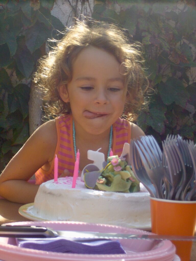 Young Lila licks her lips, staring a a colorful cake with candles on top. There are leafs on the wall behind her and some forks in a cup on the table she is next to.