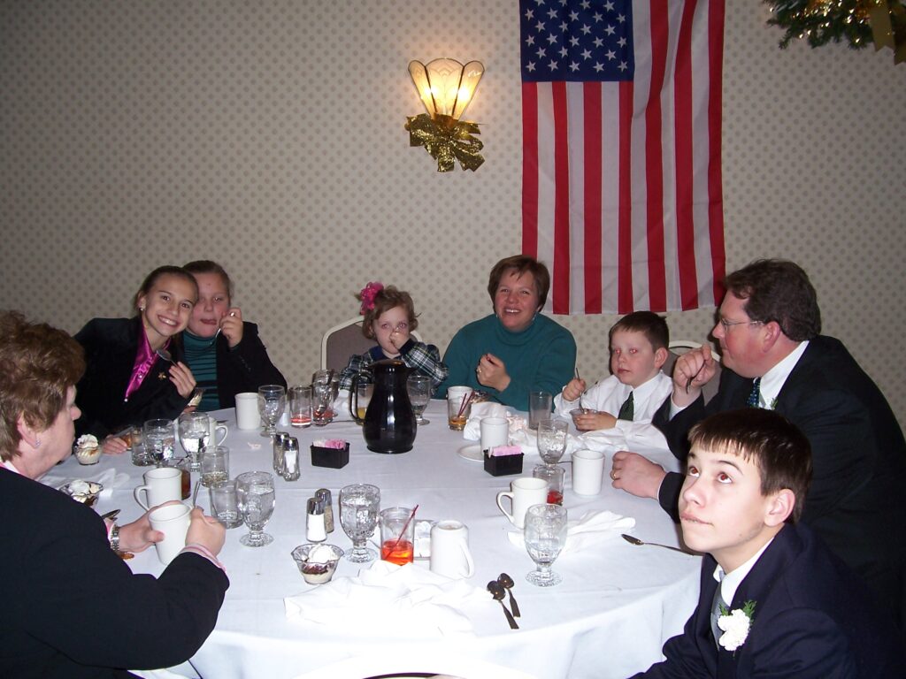 A young post sits at the center of the table with a pink bow and a spoonful of ice cream. Their cousins and siblings sit around the table with their parents. Photo was taken at a family funeral, but often this crowd would spend holidays together.