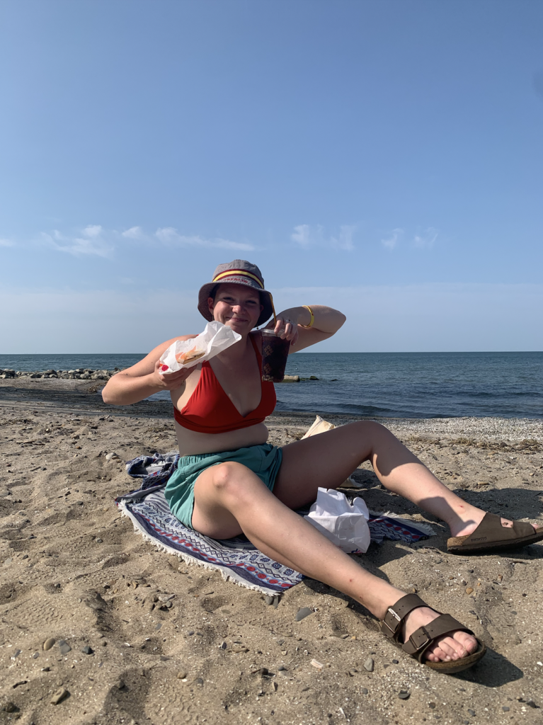 A photo of Post sitting on the beach of Lake Erie during Fall of 2020. They’re in a red swimsuit top, blue shorts, and rocking an Oberlin College floppy hat. In their hands are an iced coffee from Dunkin and a plain bagel with cream cheese. They’re smizing and are sunburnt. 
