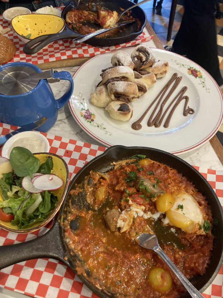 On the table there are two sets of Shakshuka, one plate of salad, one teapot, and one plate of Nutella cinnamon rolls.