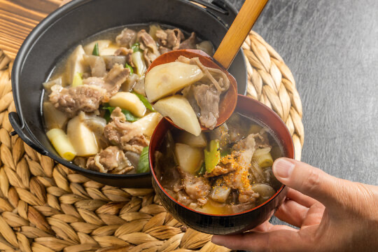 A bowl being filled with Imoni from a larger pot, the ladle full of taro potatoes and a thin slice of pork.